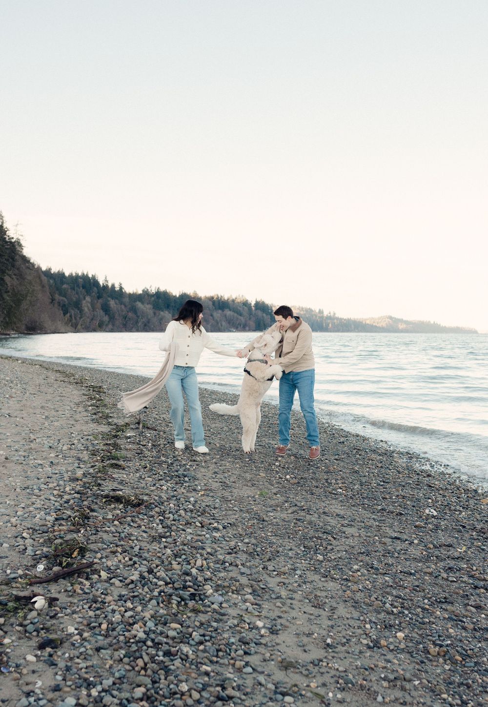 Three people strolling together on a rocky beach at sunset with evergreen trees in the background.