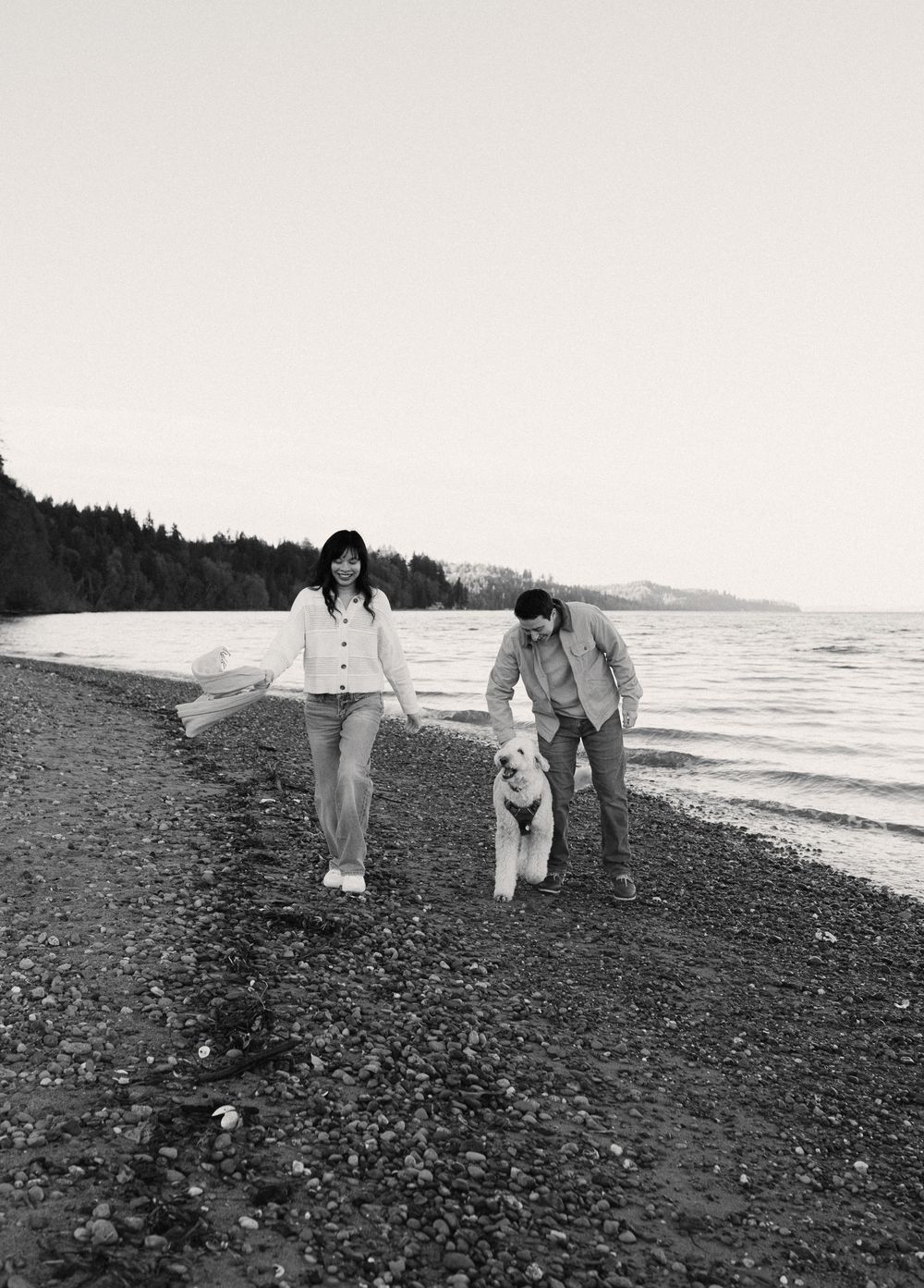 Black and white photo of couple walking dog along rocky beach shoreline.
