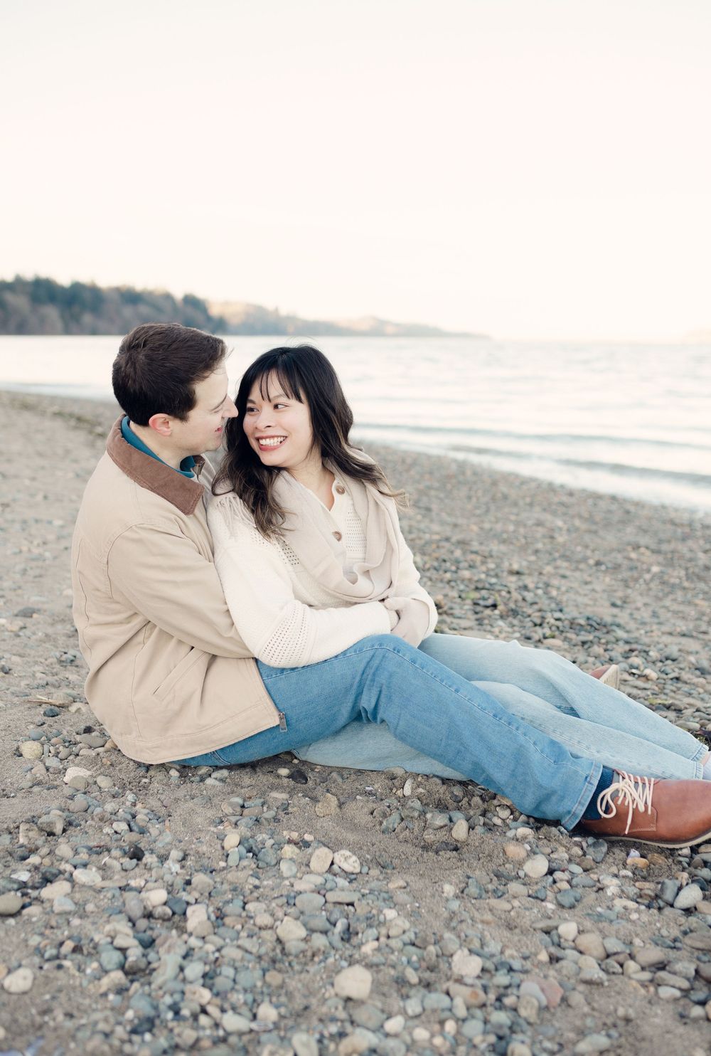 A couple sits together on a rocky beach at sunset, dressed casually in jeans and light colored sweaters.