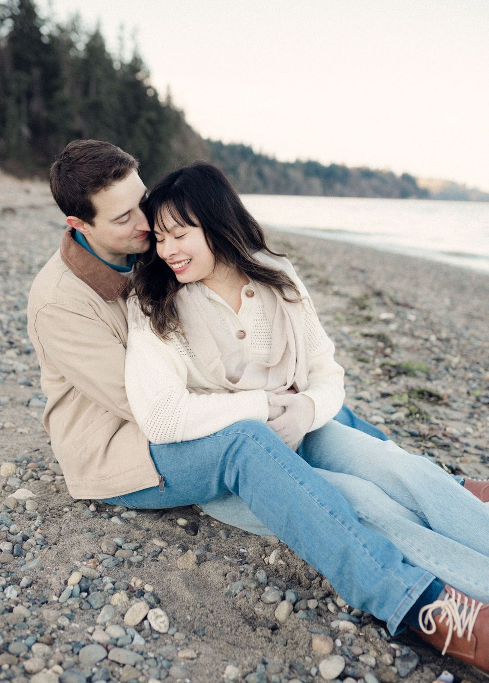 A couple in light colored sweaters and jeans sits together on a pebble beach near the water.
