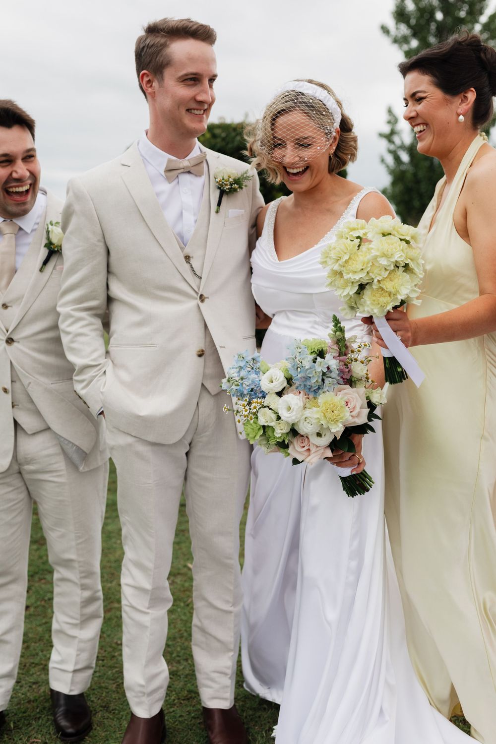 A wedding party shares a joyful moment outdoors, with the wedding party wearing light colored attire and holding pastel bouquets.