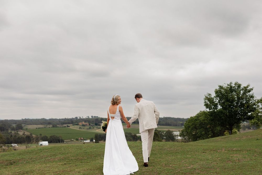 A couple in wedding attire walks hand in hand across a scenic hilltop meadow with dramatic cloudy skies overhead.