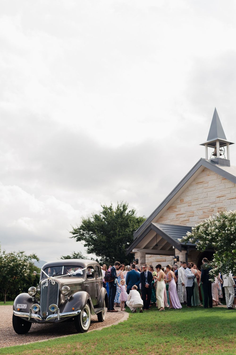 Wedding guests gather outside a charming white chapel with a vintage car parked in front on a cloudy day.