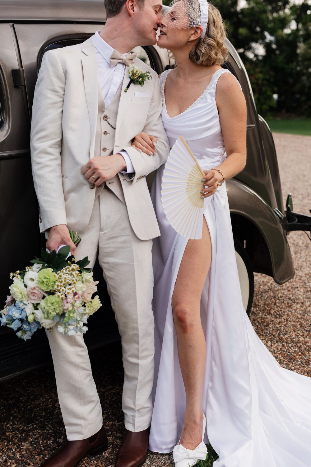 A romantic wedding couple in white attire poses next to a vintage car in a garden setting.