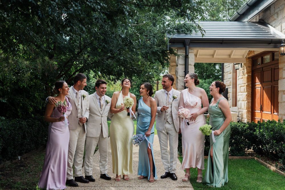 Wedding party members in pastel dresses and khaki suits pose together on a lush green lawn.