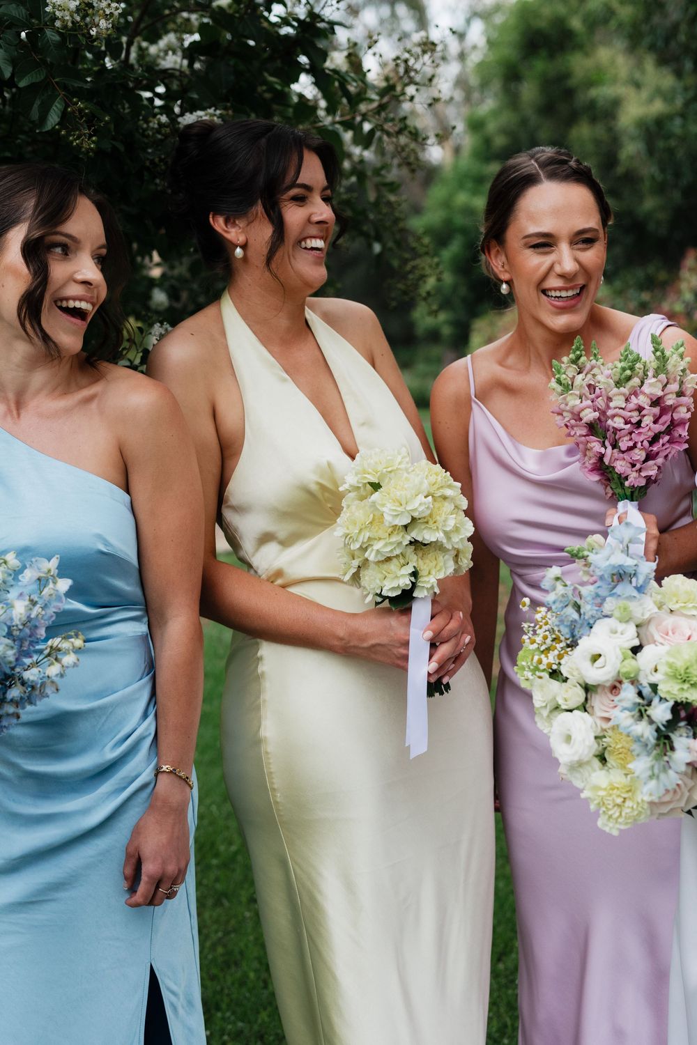 Bridesmaids in pastel dresses holding colorful bouquets laugh together outdoors.