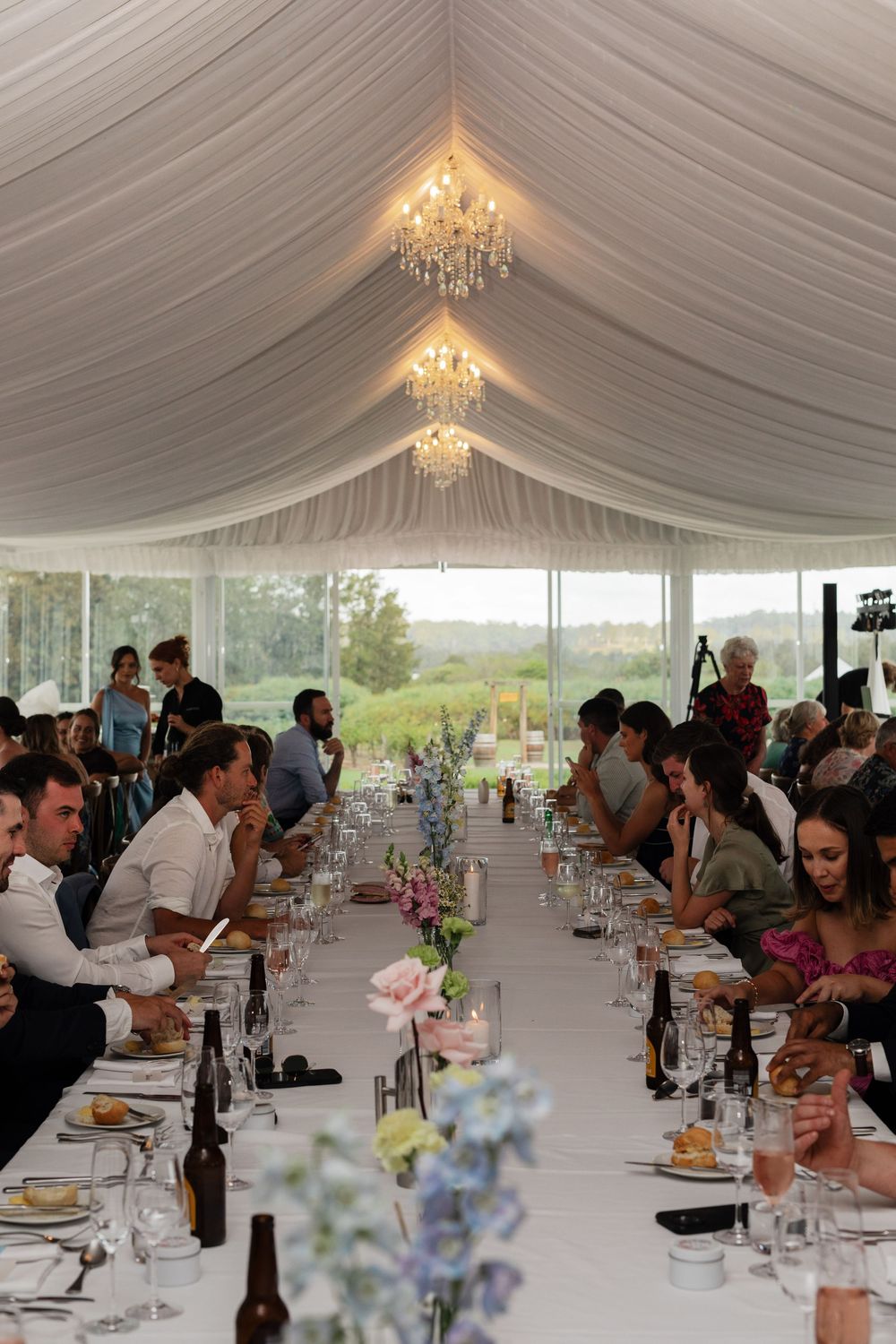 Wedding reception under a white tent with elegant chandeliers and long tables set for dinner service.