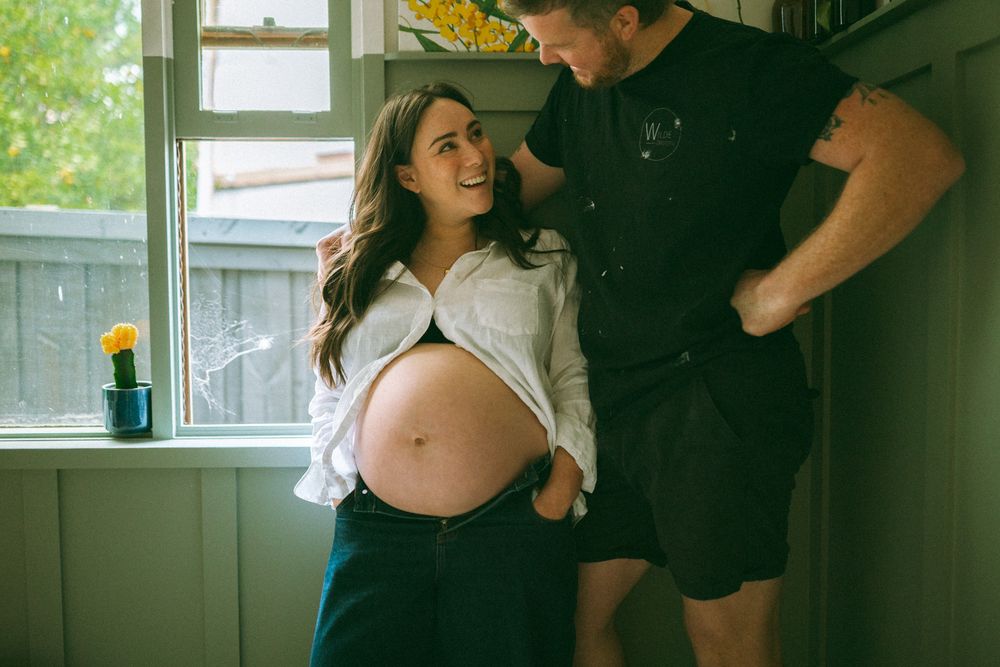 A pregnant woman in a white blouse stands by a window while interacting with someone in dark clothing.