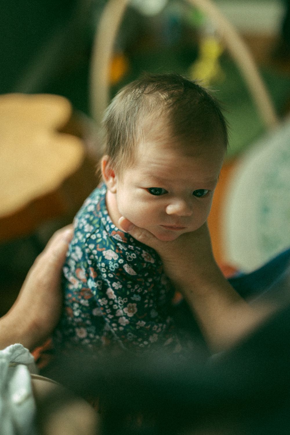 A small baby wearing a floral patterned dress looks up at the camera with an inquisitive expression.