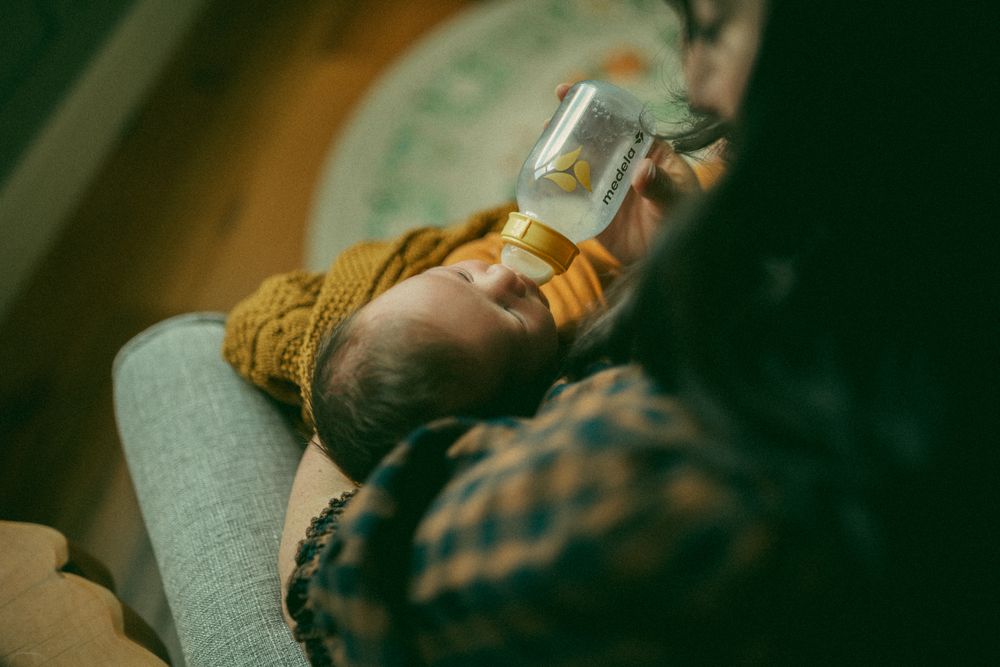 A baby in a yellow sweater lies on a gray couch while drinking from a bottle in moody lighting.