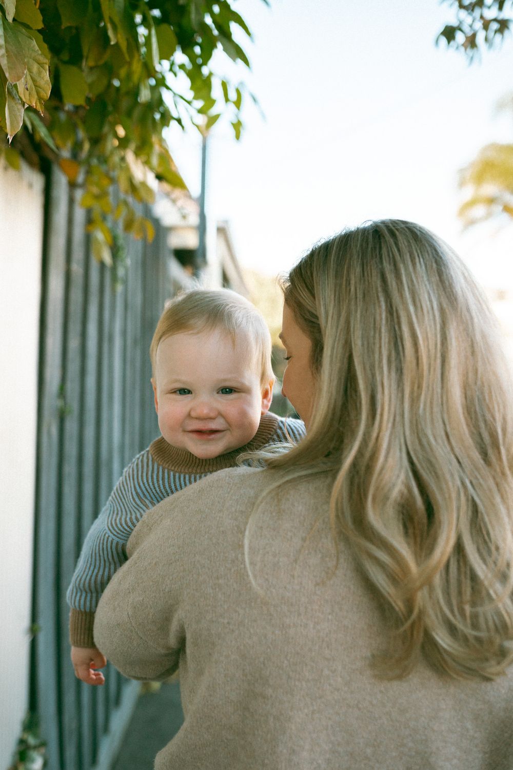 A parent and child in coordinating beige and gray sweaters share a tender moment outdoors near greenery.