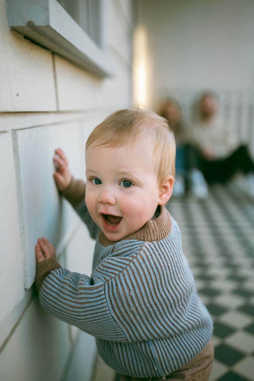 A small child in a gray sweater crawls across a black and white checkered floor.