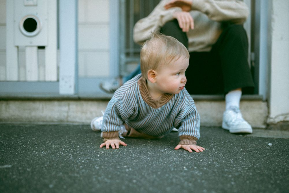 Baby in gray sweater crawls on ground while someone stands nearby in white sneakers.