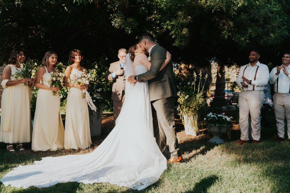 Outdoor wedding ceremony with bridal party in cream dresses and groomsmen in light suits standing on sunny lawn.
