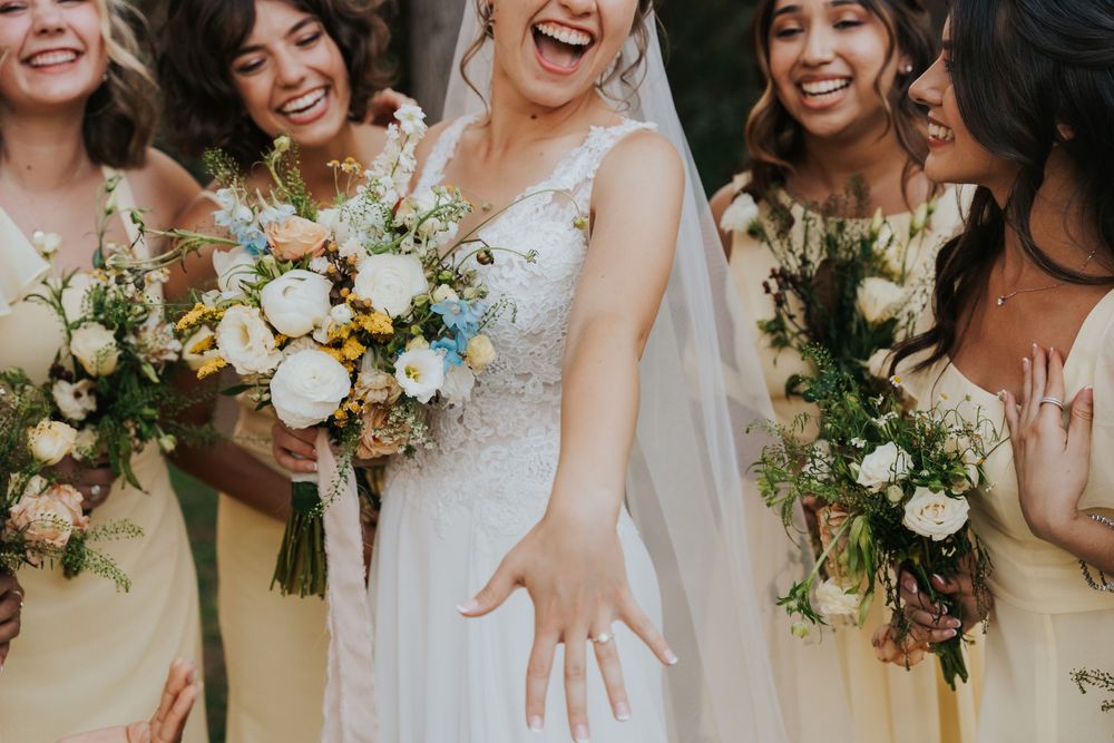 A wedding party in blush pink dresses holds elegant white and green floral bouquets while laughing together.