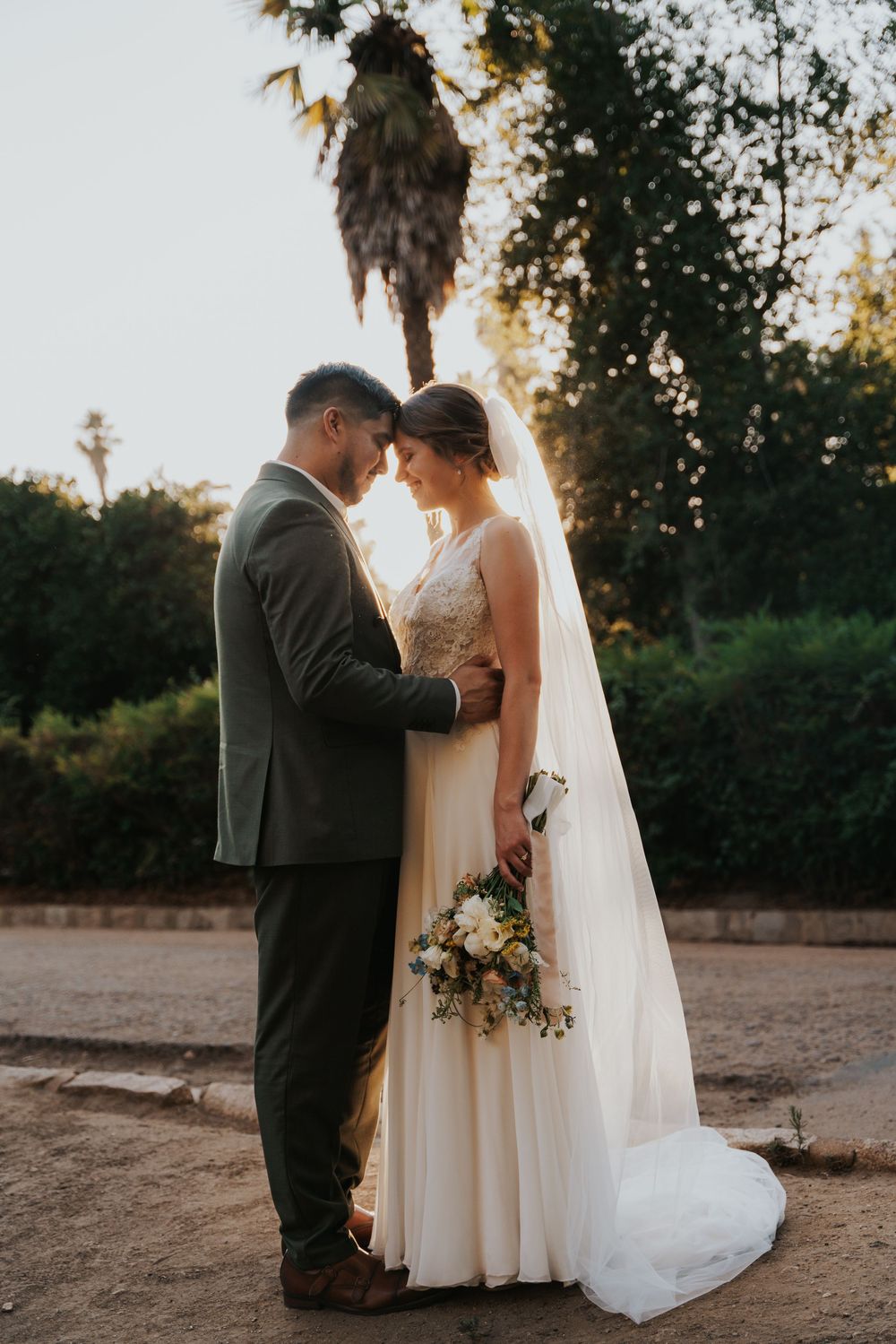 A romantic wedding couple shares an intimate moment at sunset with a palm tree and natural backdrop.