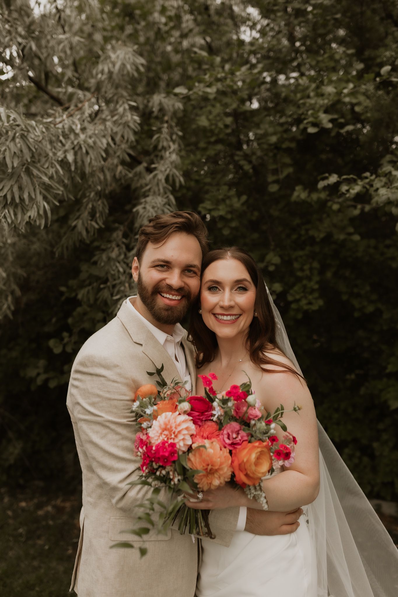 Newlyweds pose with bright coral and pink bouquet against natural outdoor backdrop.