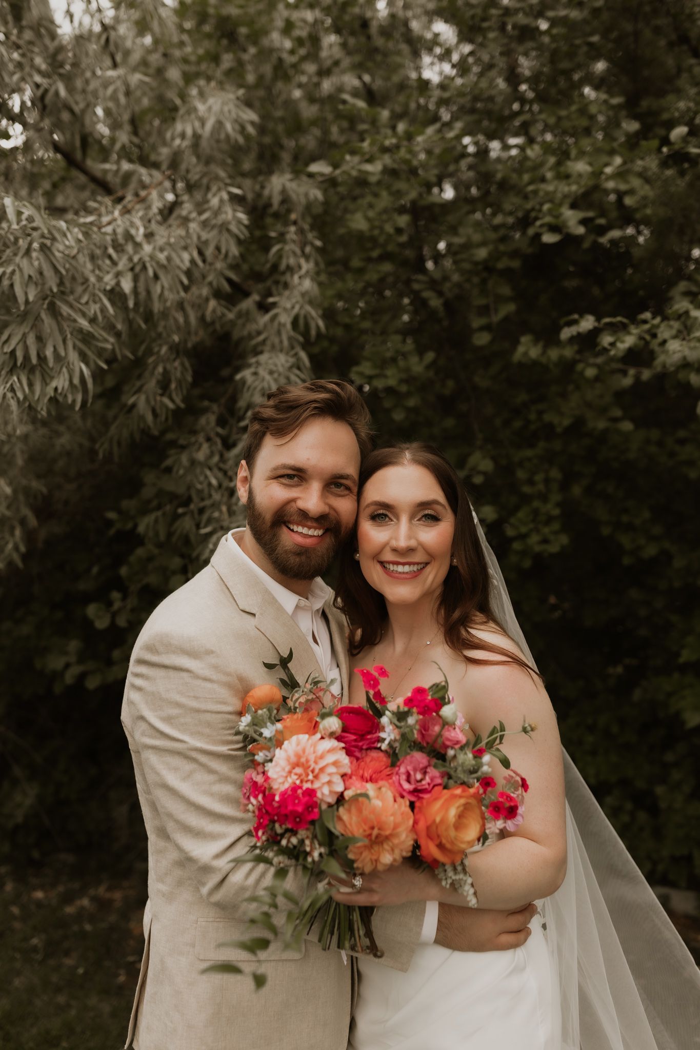 A wedding couple in a garden setting with the bride holding a vibrant pink and coral bouquet.