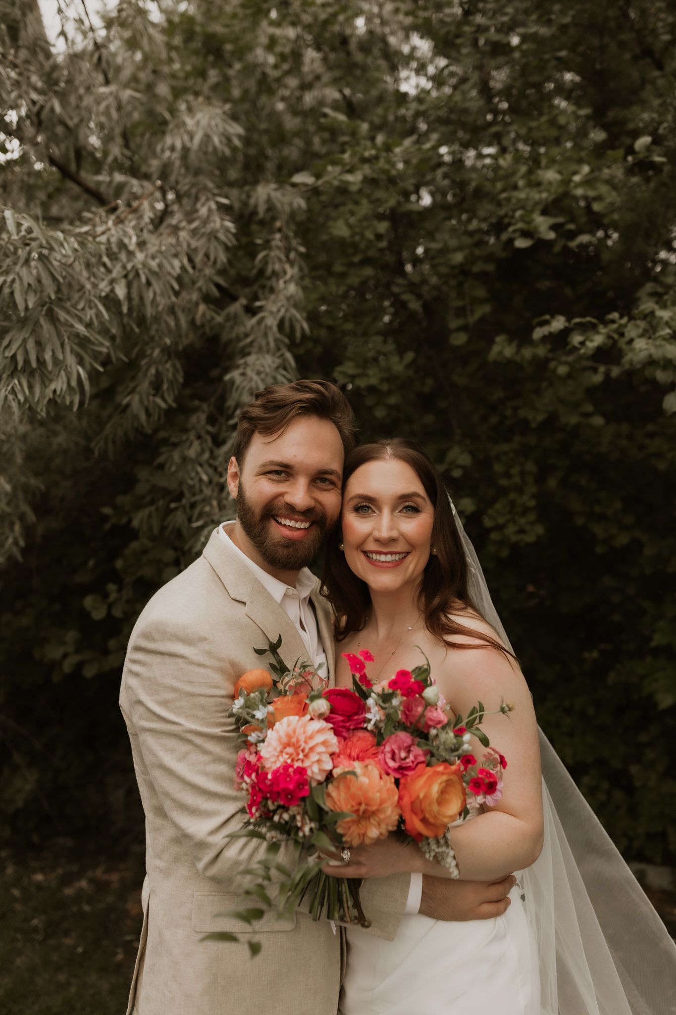 Newlyweds embrace outdoors while holding a colorful bouquet of pink and orange flowers.