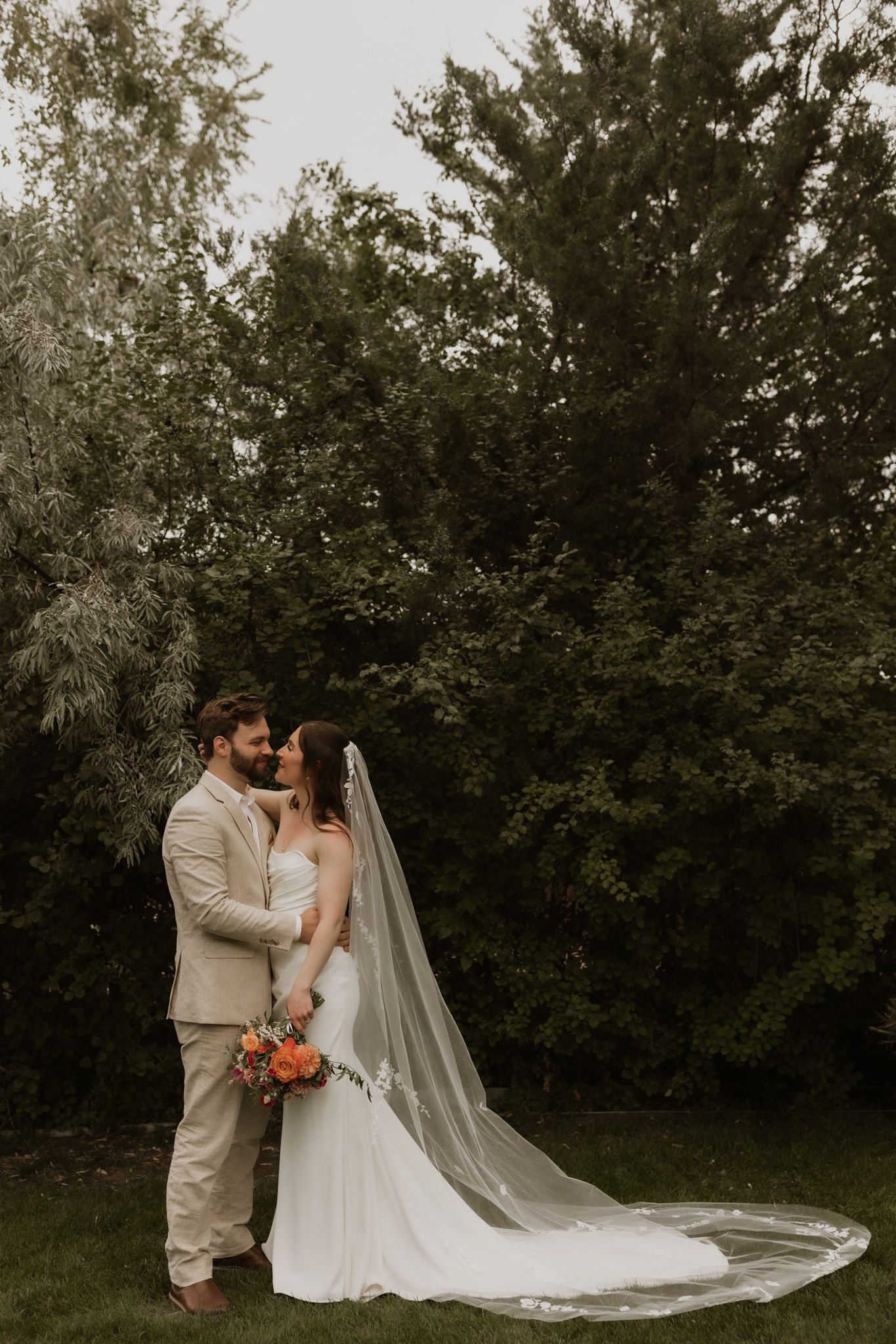 A romantic wedding portrait with a long flowing veil against a backdrop of evergreen trees.