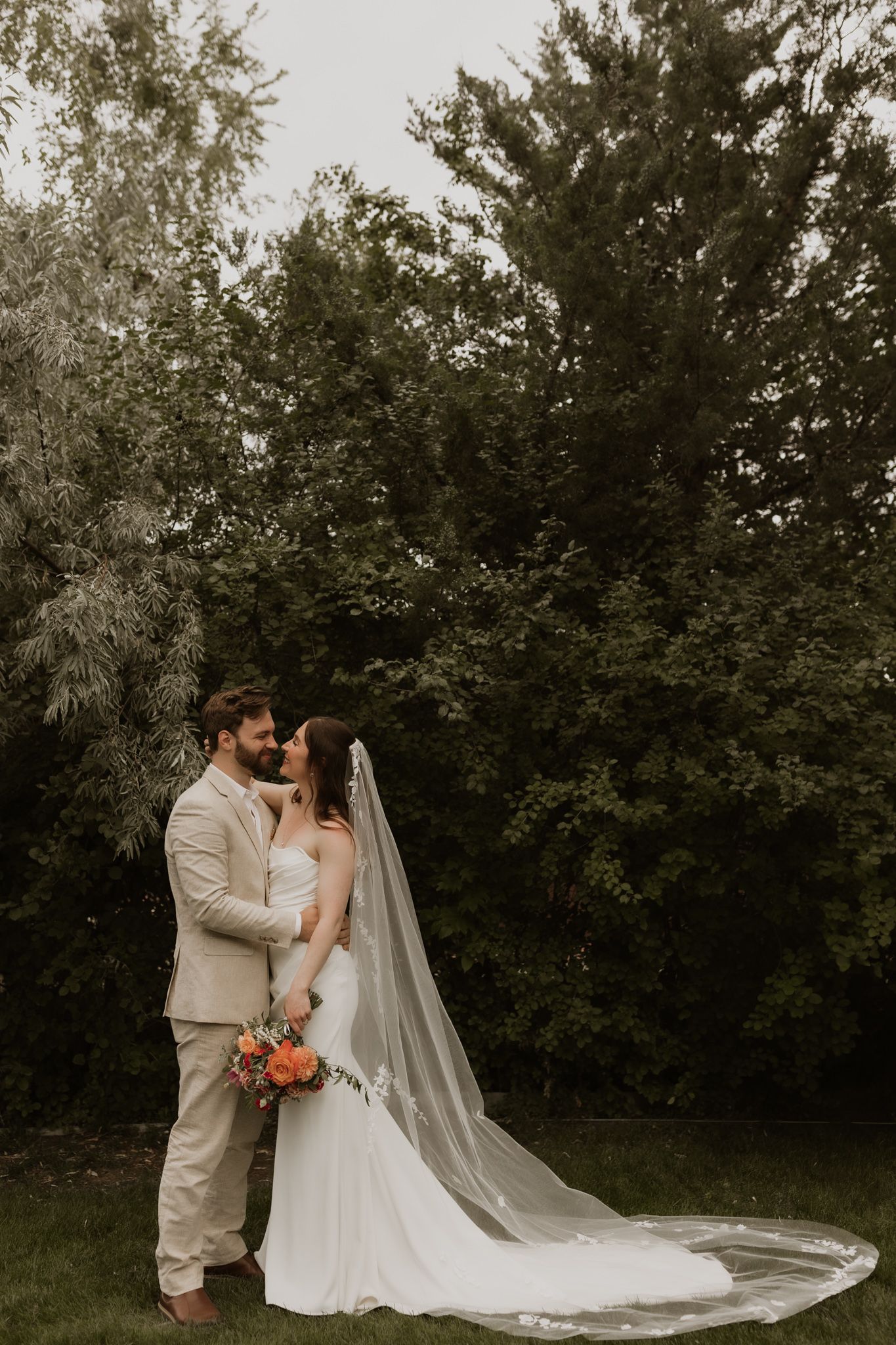 A romantic wedding couple shares an intimate moment in a garden setting with a flowing white veil trailing behind.
