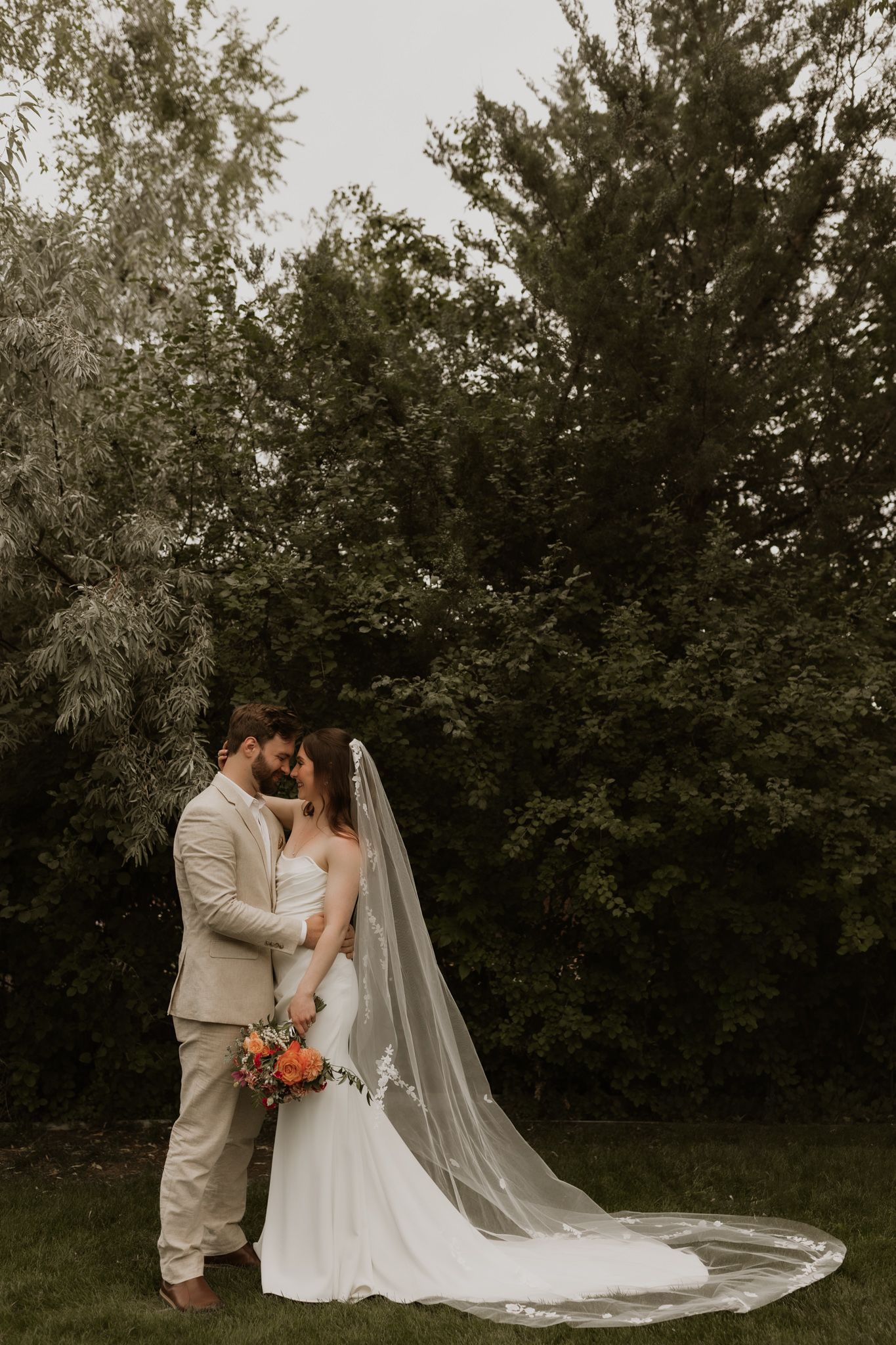 An intimate wedding moment captured in a garden setting with a flowing white veil and beige suit.