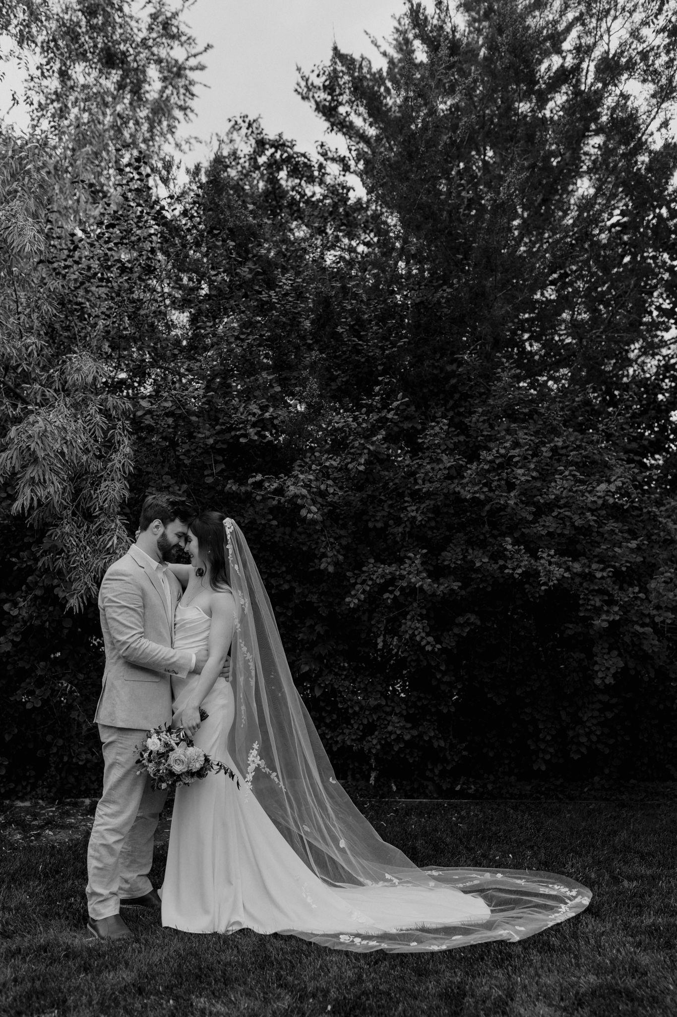 A black and white wedding portrait showing a couple in a natural outdoor setting with flowing veil.