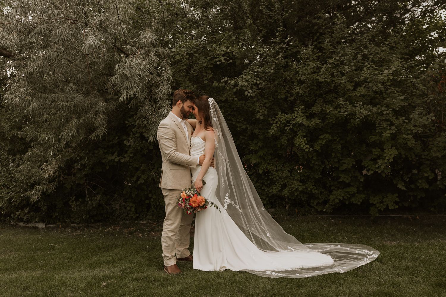 An intimate wedding portrait with the bride's long veil trailing on grass in a garden setting.