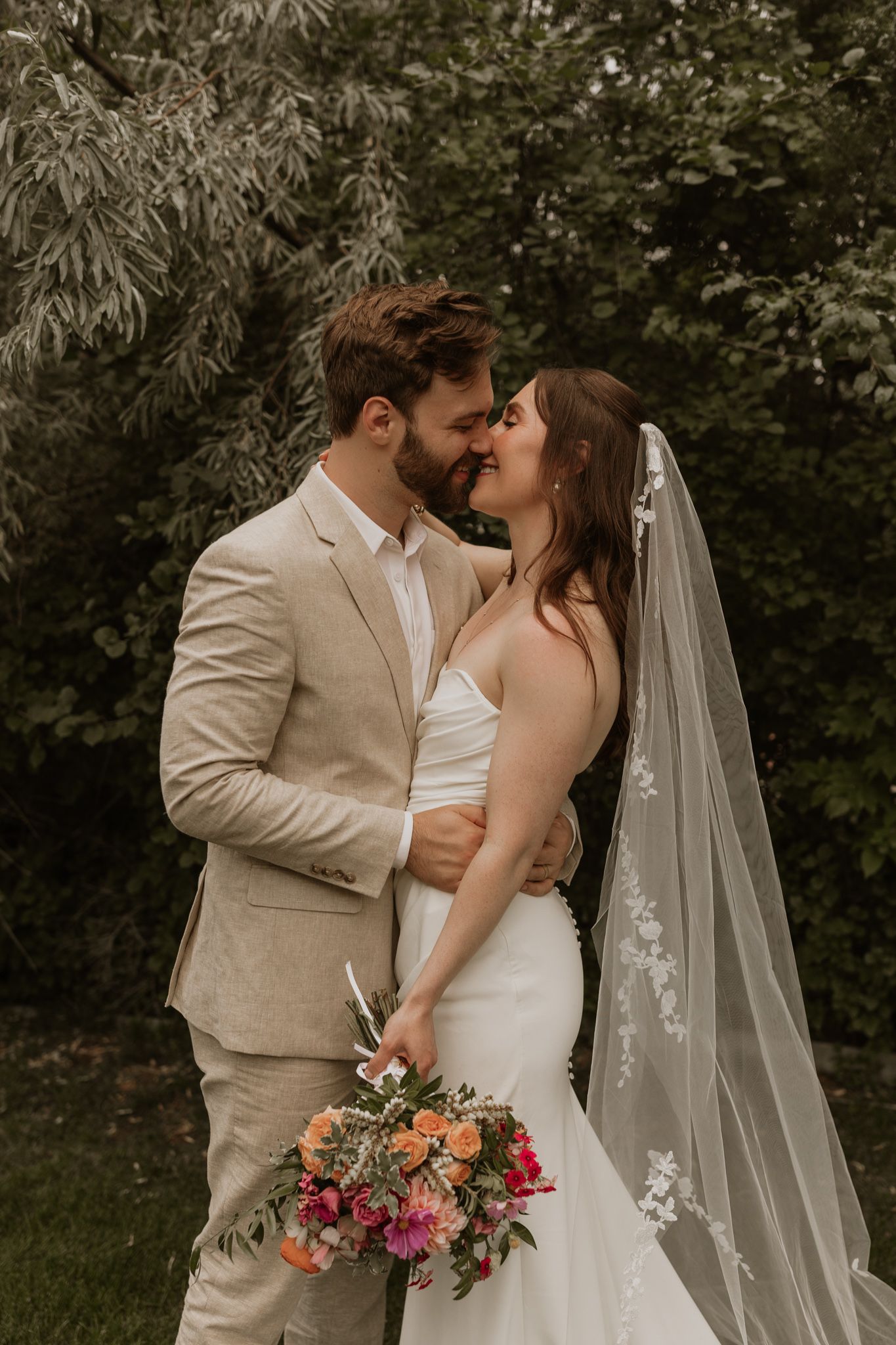 A couple shares a tender moment in their wedding attire with colorful bouquet against natural backdrop.