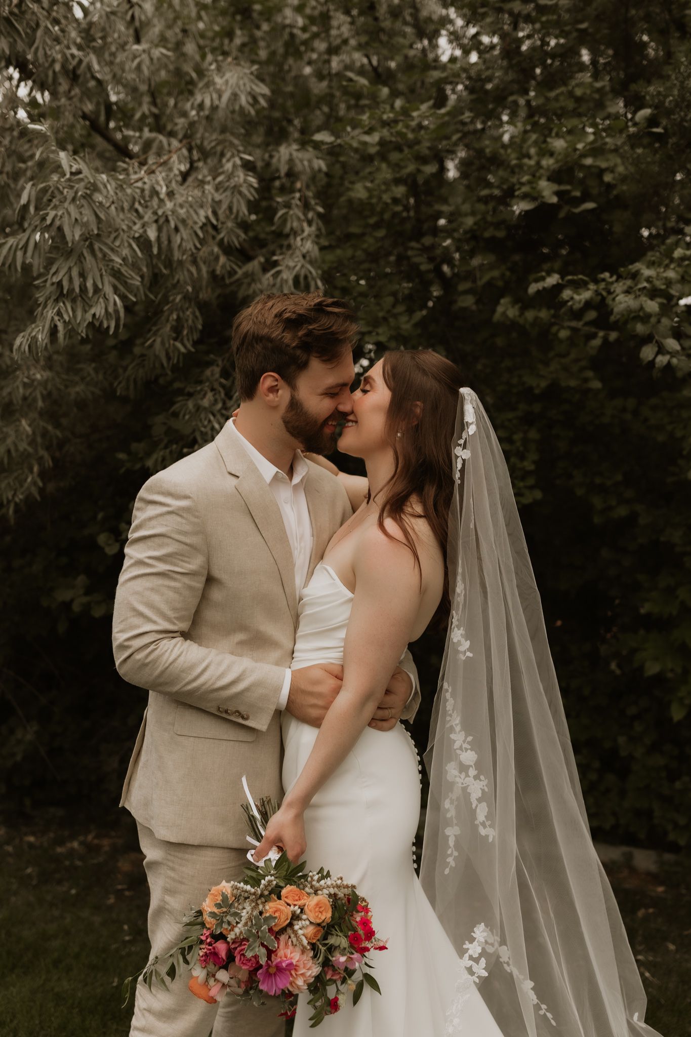 A romantic wedding couple shares intimate moments in a garden setting with a flowing veil and colorful bouquet.