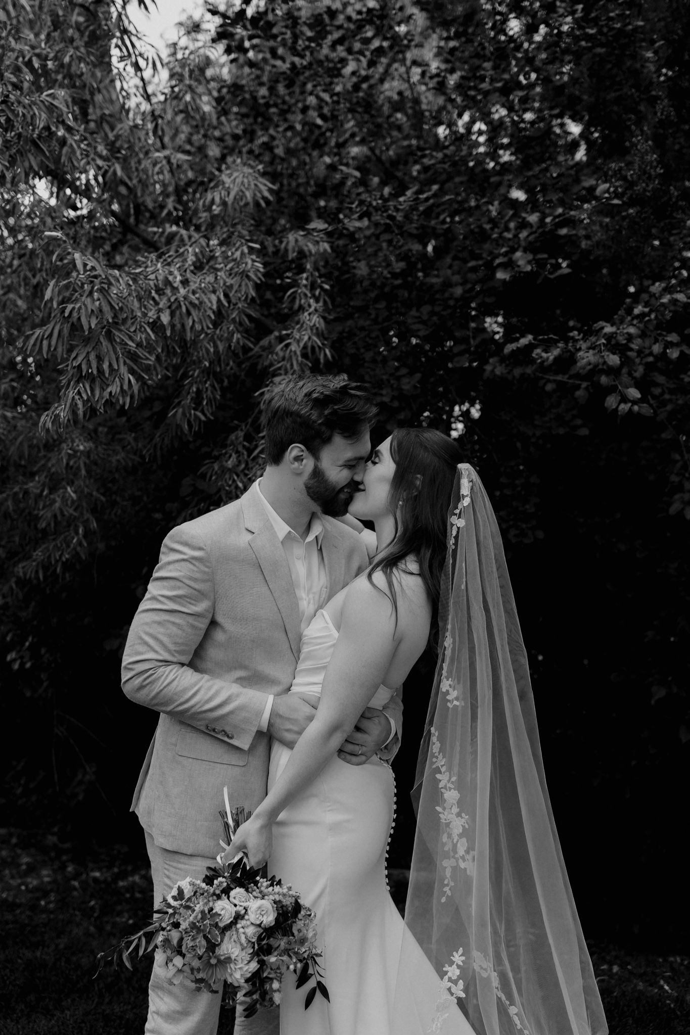 A romantic black and white photo series of newlyweds embracing in a garden setting with a flowing veil.