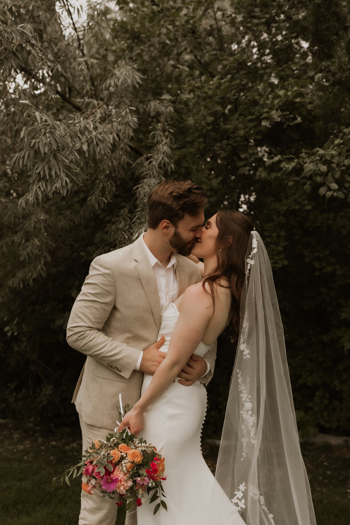 Romantic wedding kiss captured in a garden setting with flowing veil and floral bouquet.