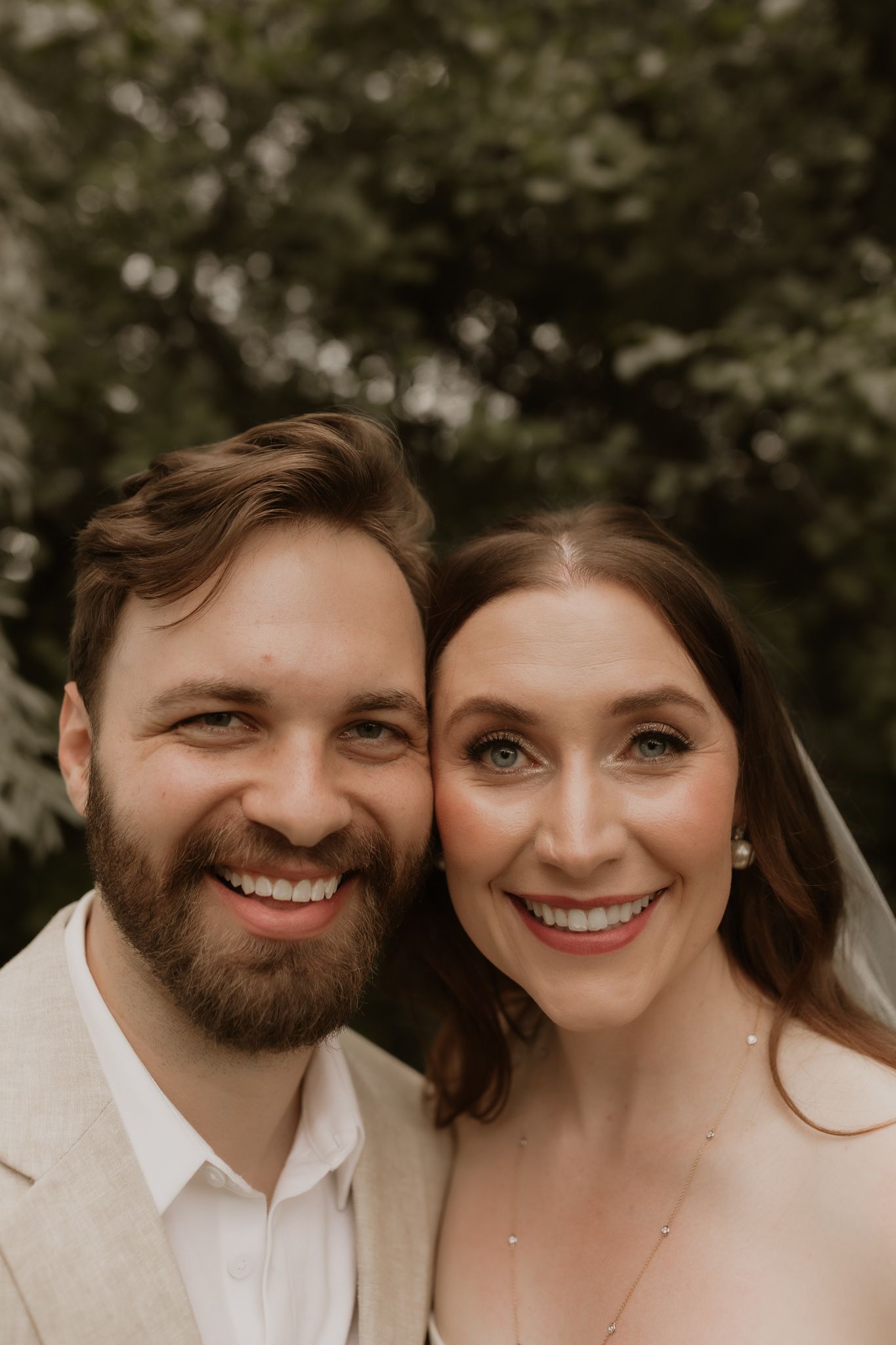 A close-up portrait of a newlywed couple sharing joyful smiles against a natural outdoor background.