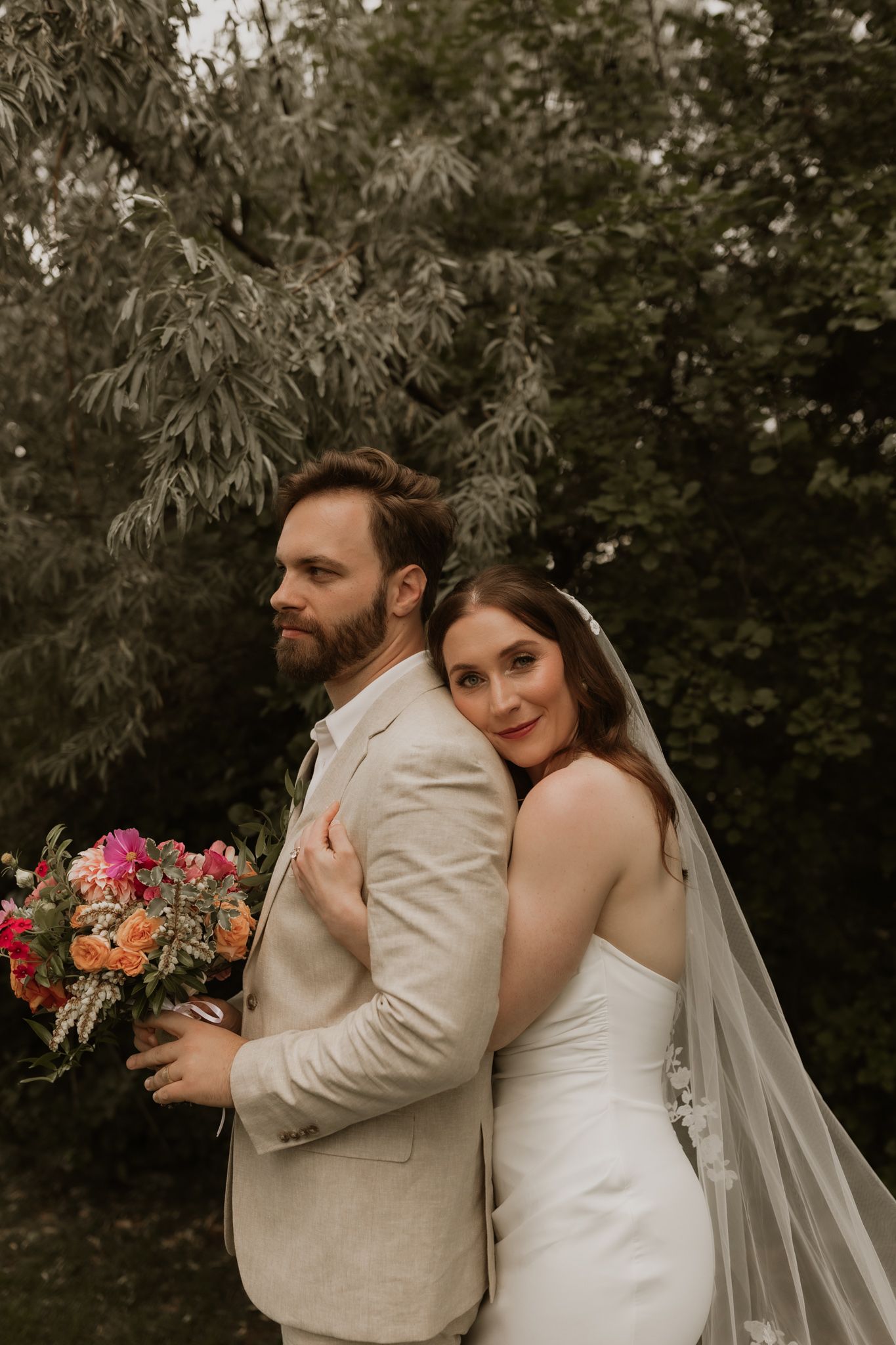 A serene outdoor wedding photo showing a couple in beige and white attire with a flowing veil.