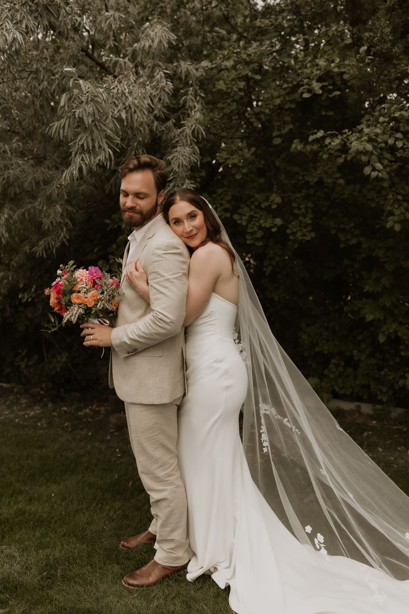 Wedding couple in beige and white attire pose together with long veil trailing behind.