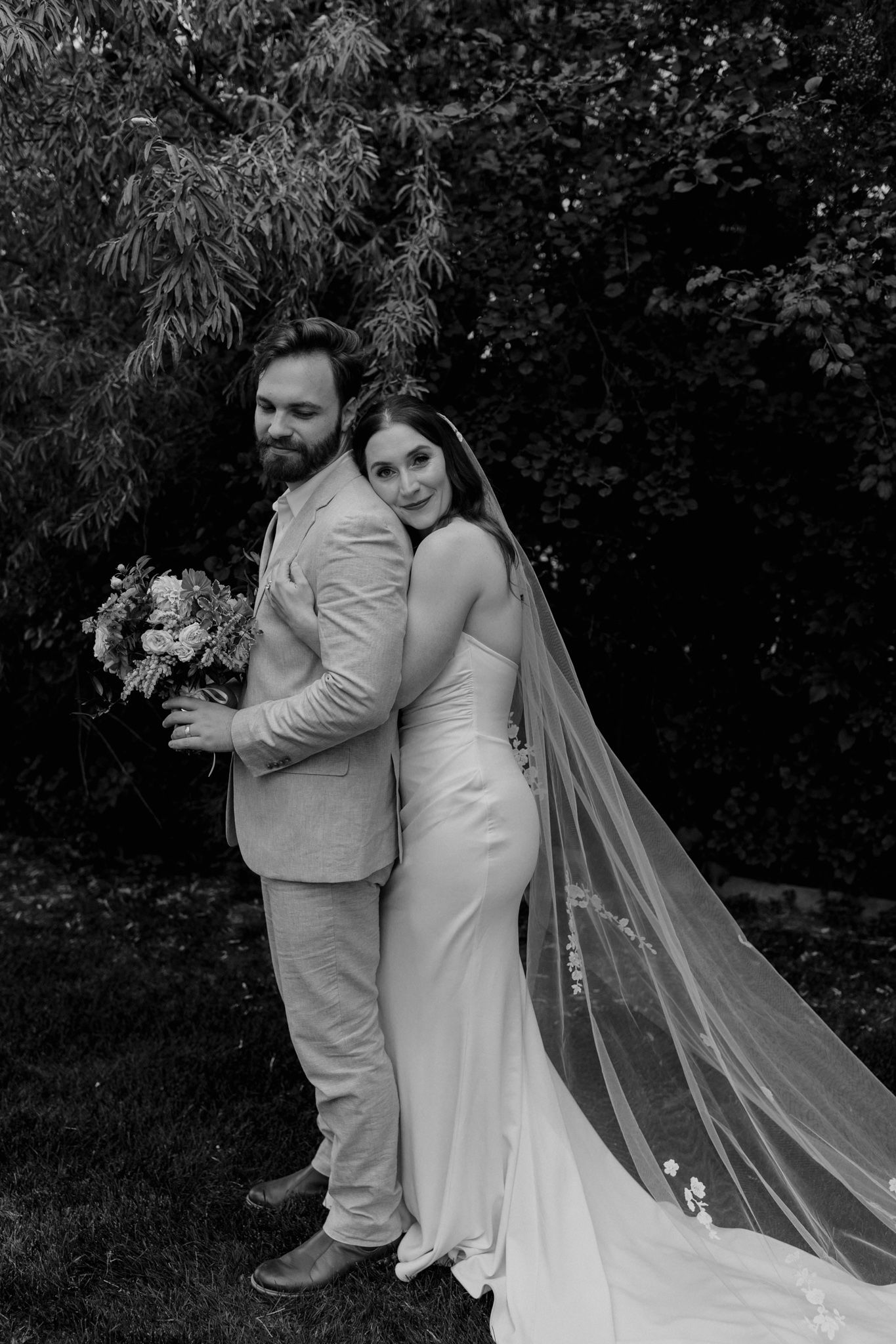 A candid black and white wedding photo showing tender moments between newlyweds in a garden.