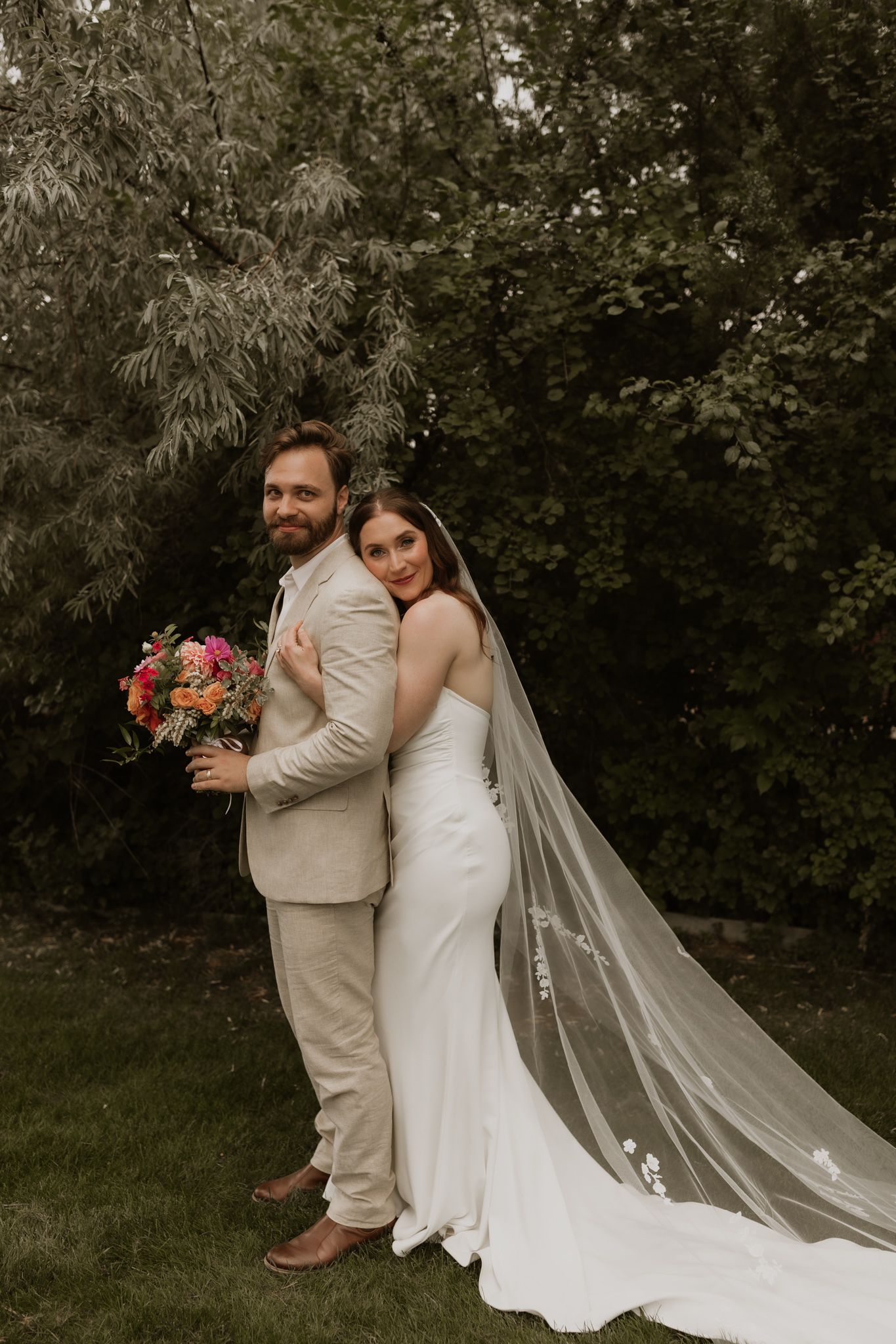 A romantic outdoor wedding photo of a couple in a garden setting with the bride wearing a flowing veil and holding a bouquet.
