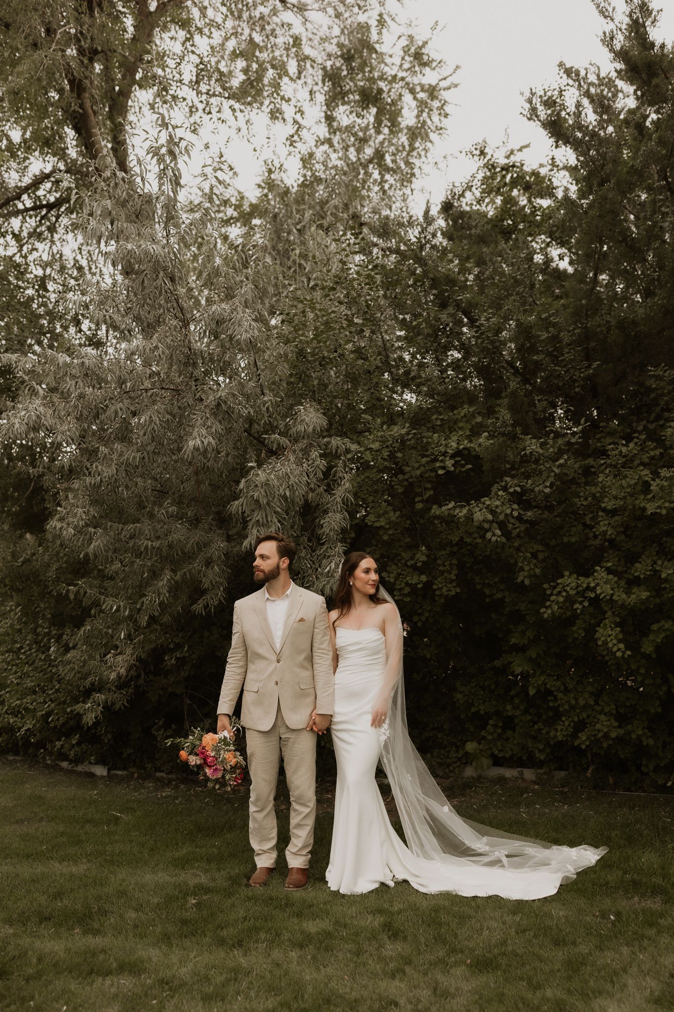Full-length wedding portrait of a couple in beige and white attire standing in a garden.
