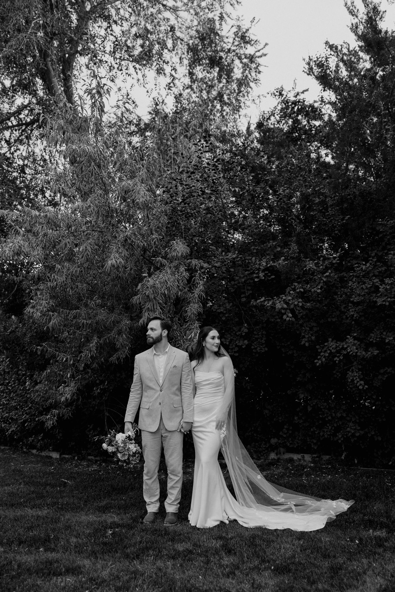 Black and white wedding portrait of a couple standing against lush garden foliage with a long trailing bridal veil.