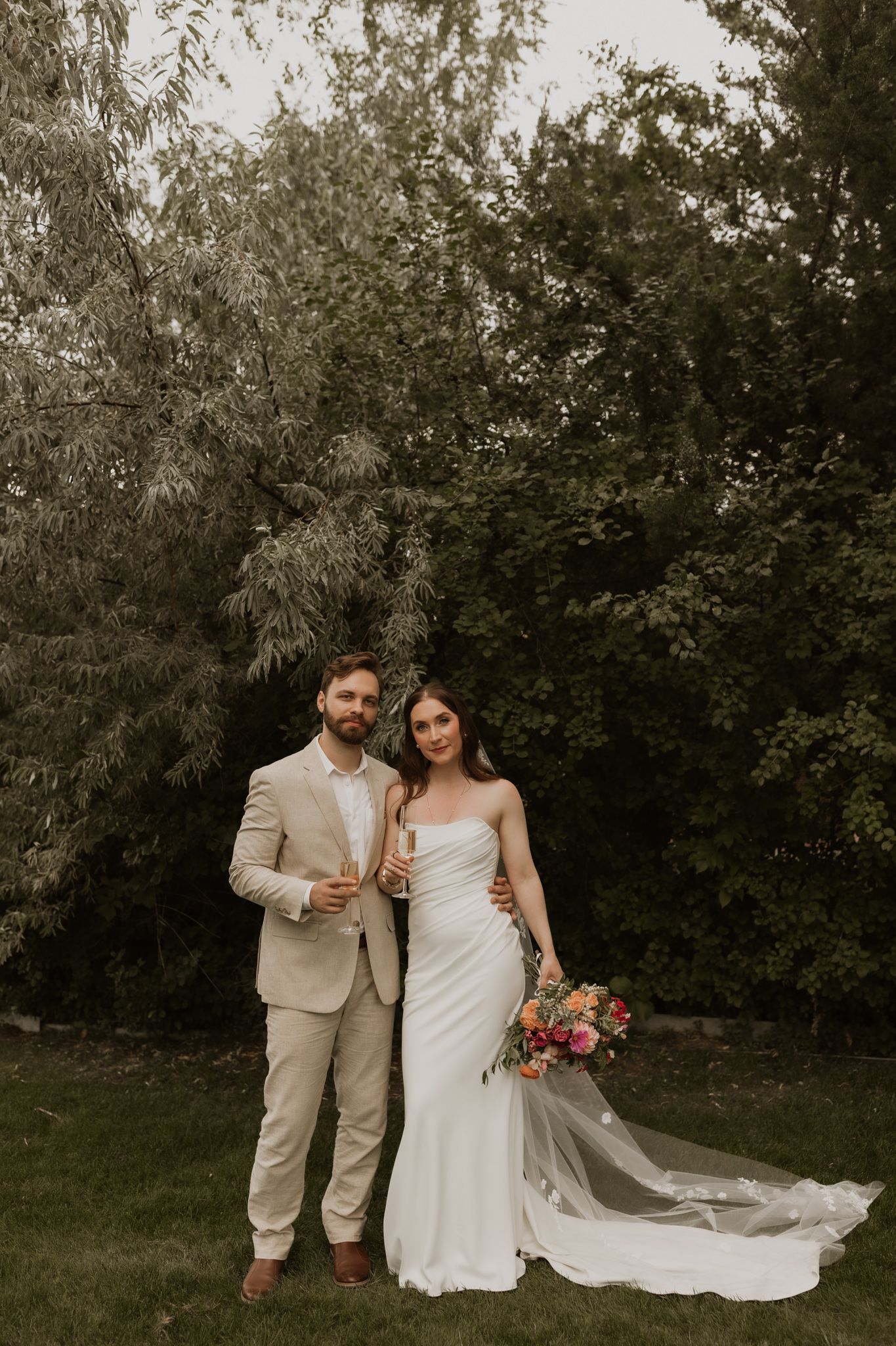 A couple in wedding attire poses together against a backdrop of lush greenery in a romantic outdoor setting.