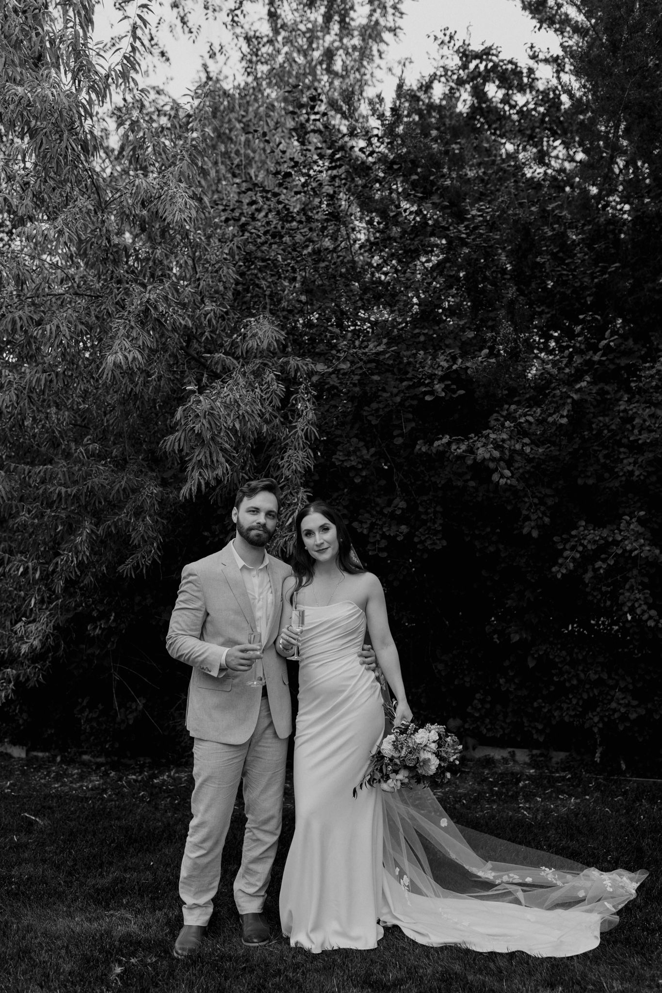 Newlyweds share a tender moment in a garden setting with the bride's flowing white dress and groom in light suit.