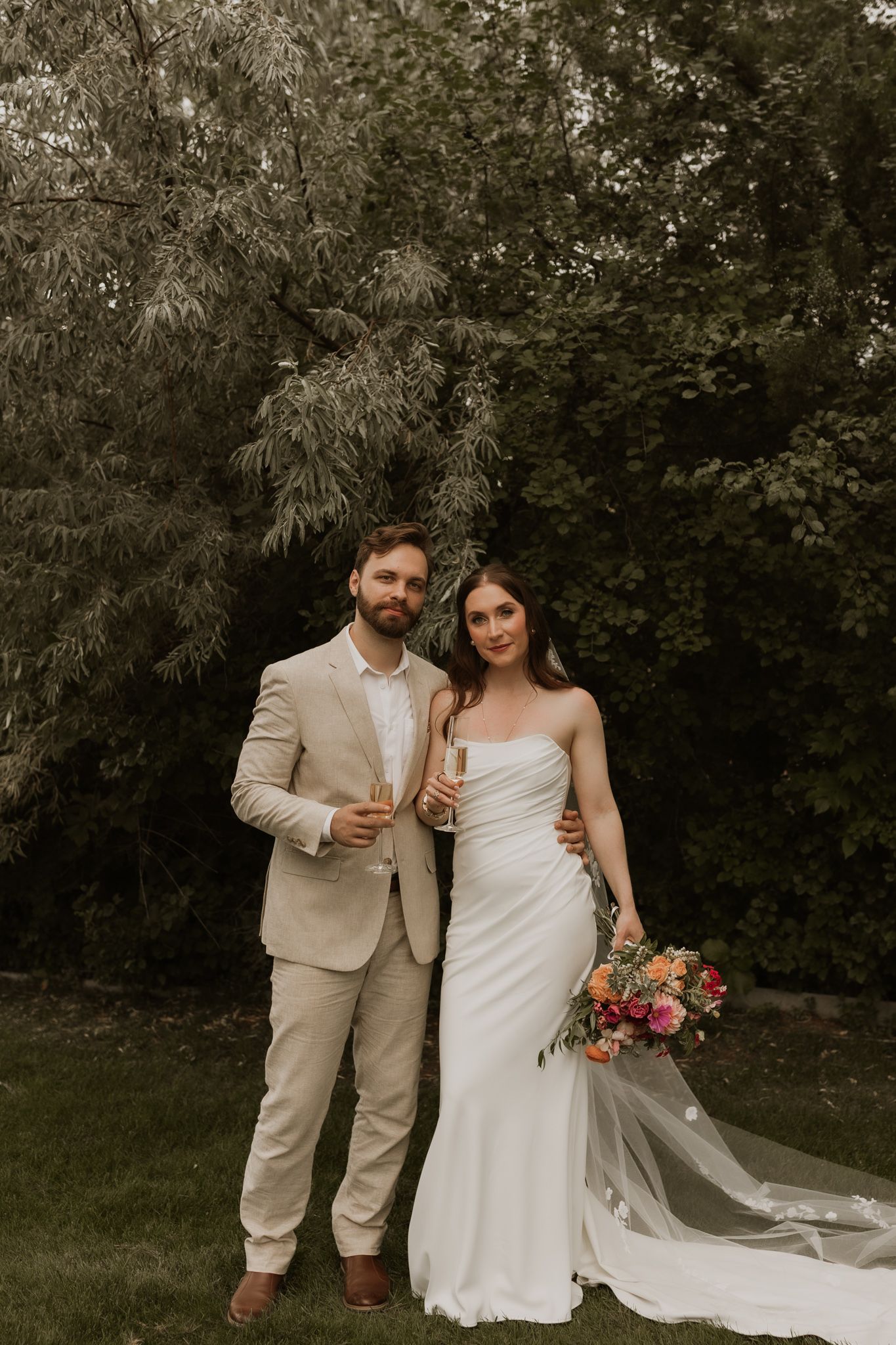 A wedding couple poses in a garden setting with elegant white dress and beige suit.