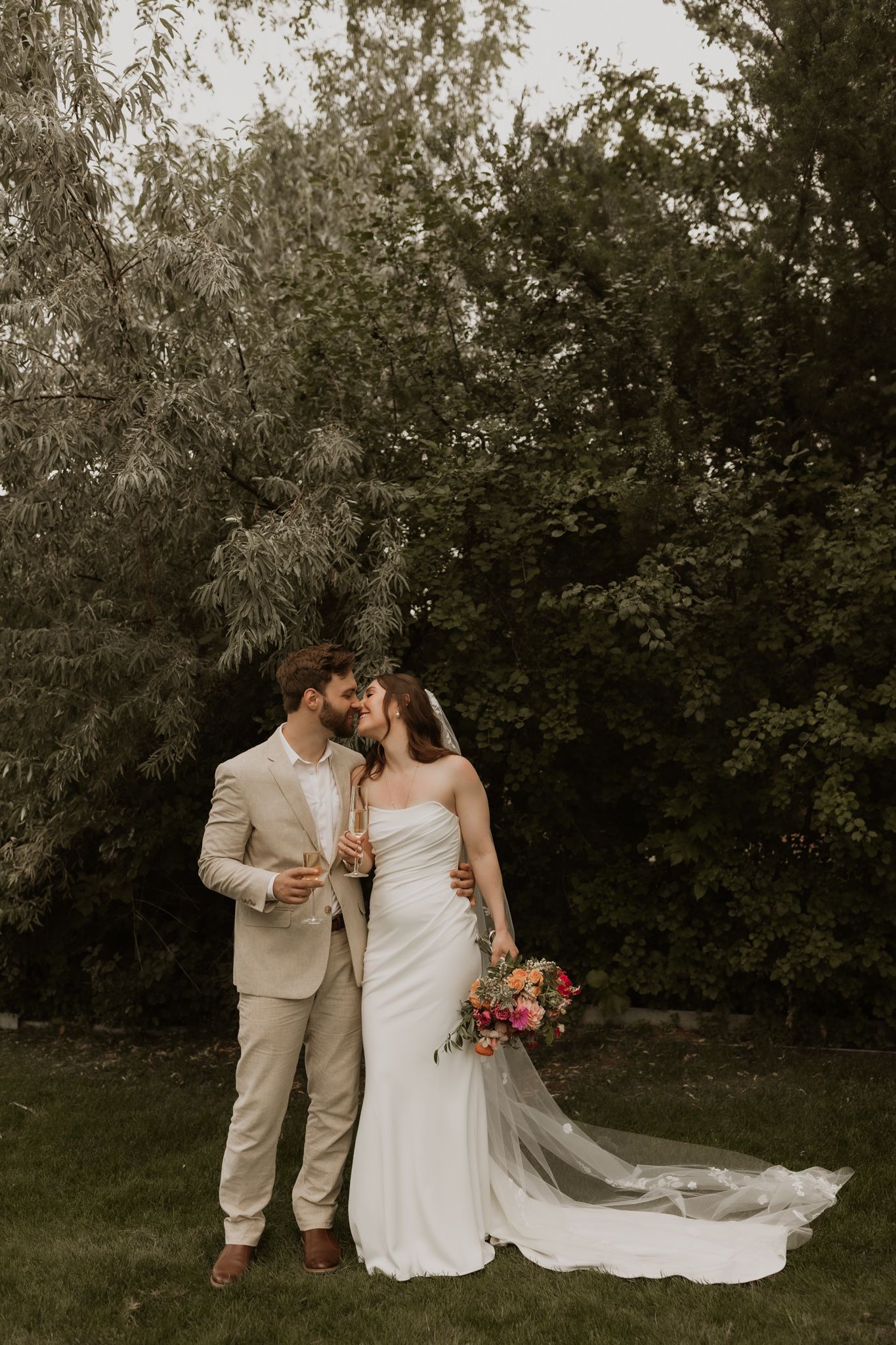 Couple shares tender moment in garden setting wearing beige suit and white wedding dress.