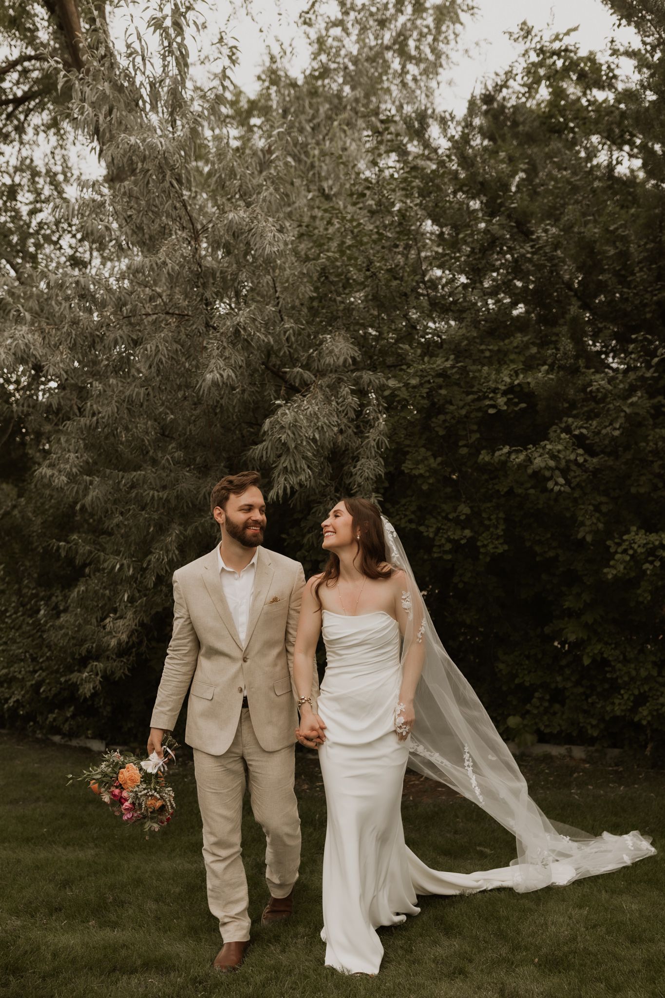 A couple in wedding attire walks together through a garden setting with flowing veil and light beige suit.