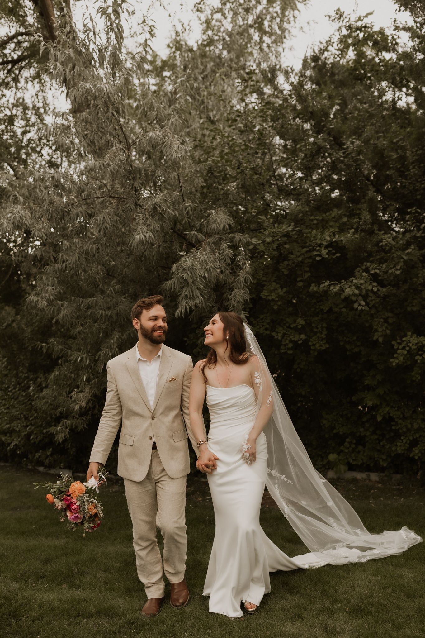 A wedding couple walks hand in hand through a garden setting with flowing veil and floral bouquet.