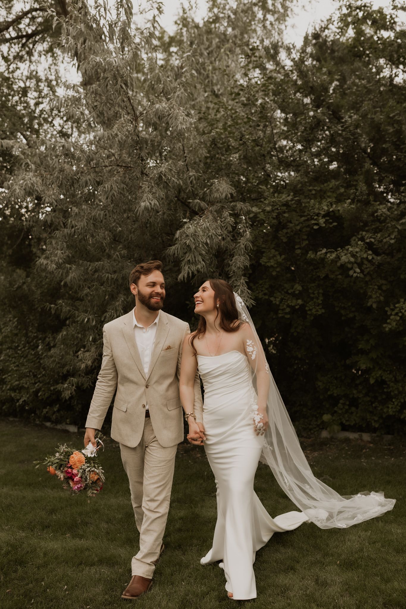 Candid walking shot of newlyweds in a garden with flowing veil and coral bouquet.