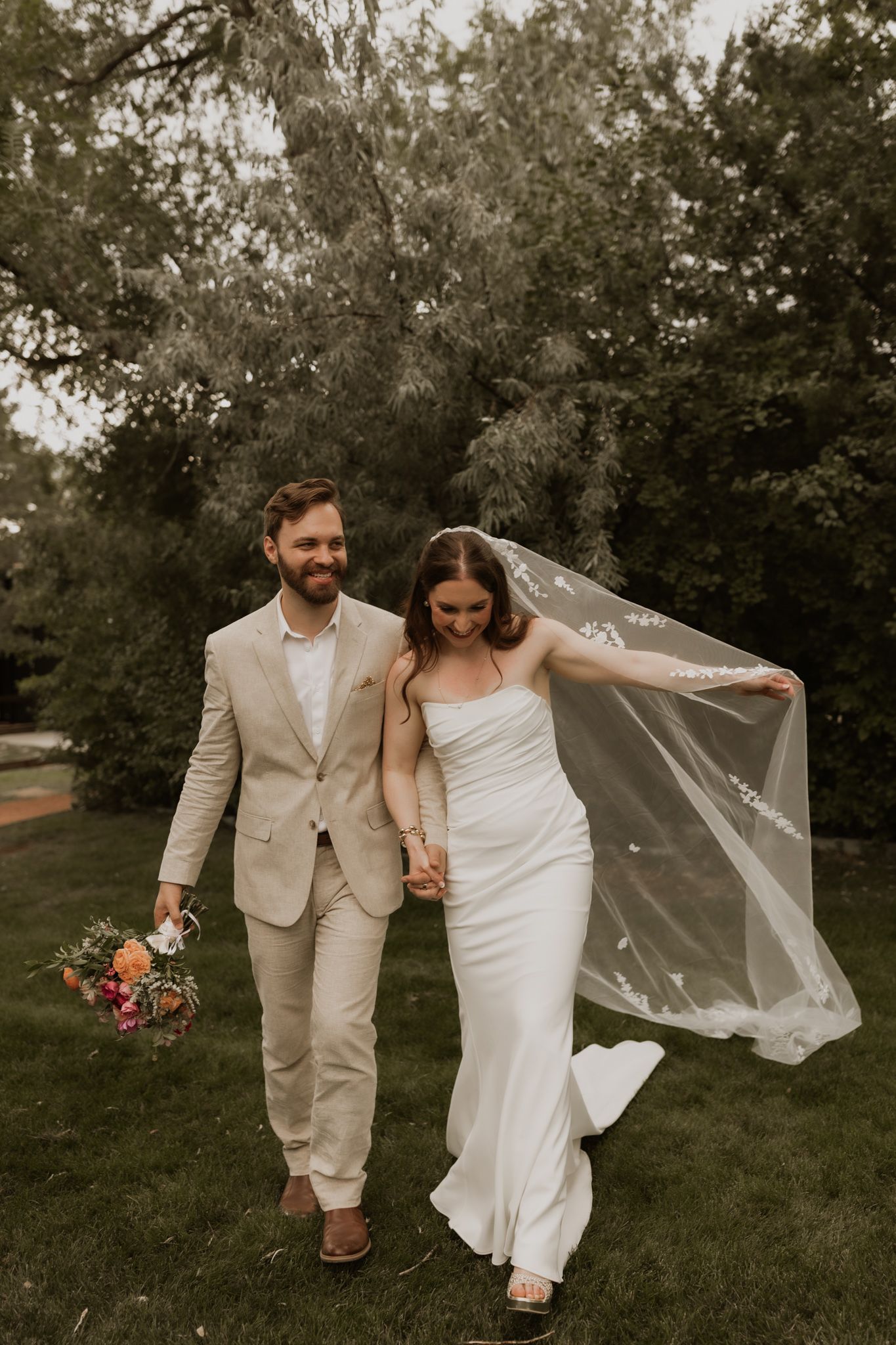 Newlywed couple walks through garden with flowing veil and beige suit on sunny day.