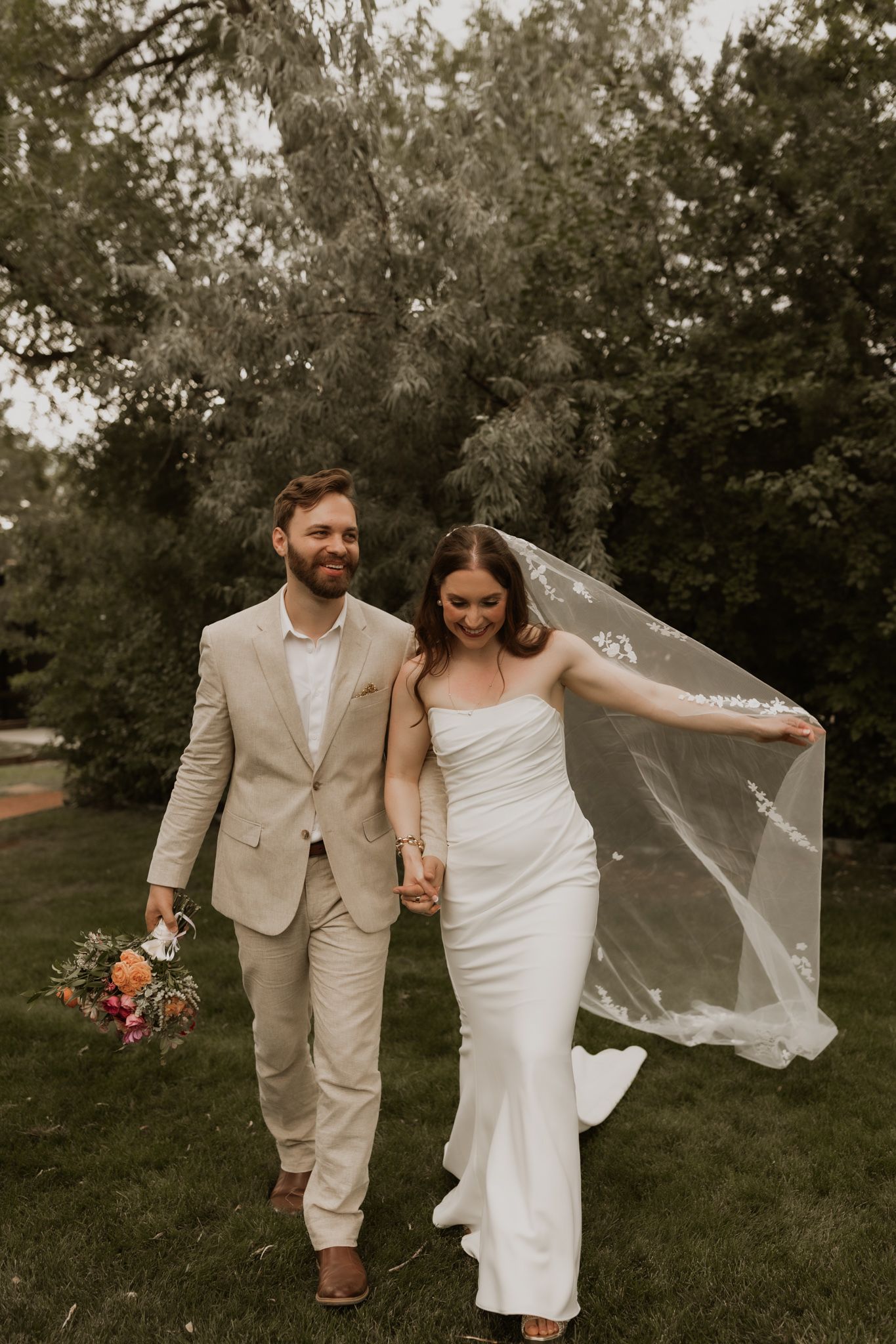 A candid wedding photo of a couple walking together outdoors with bride's veil flowing in the wind.