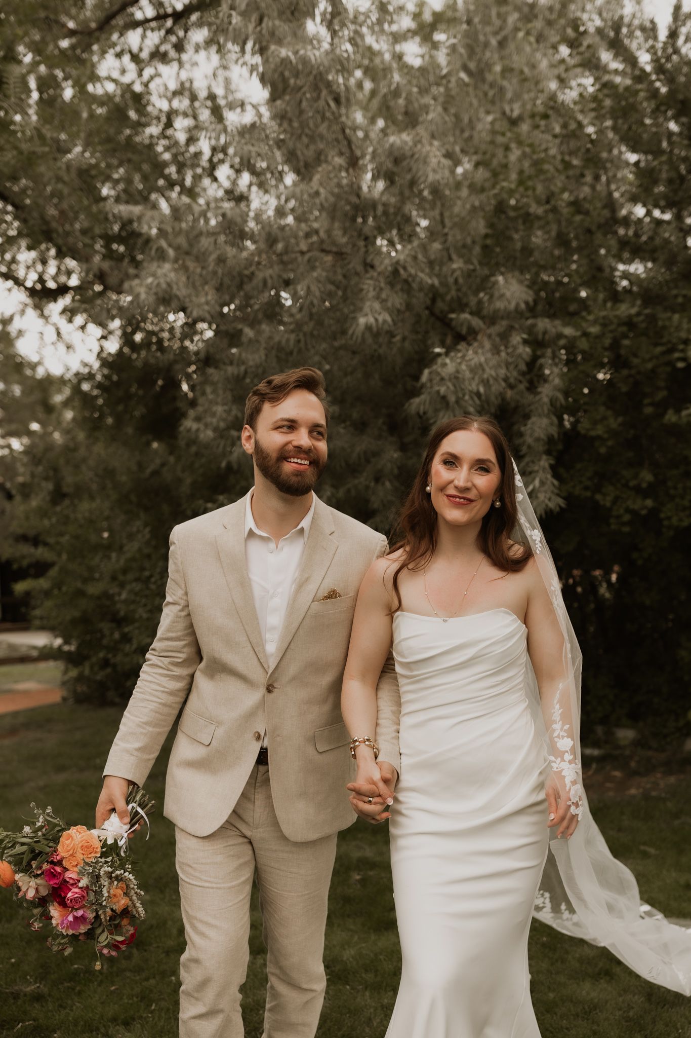 A joyful wedding portrait featuring a couple in light-colored attire holding bright floral arrangements.