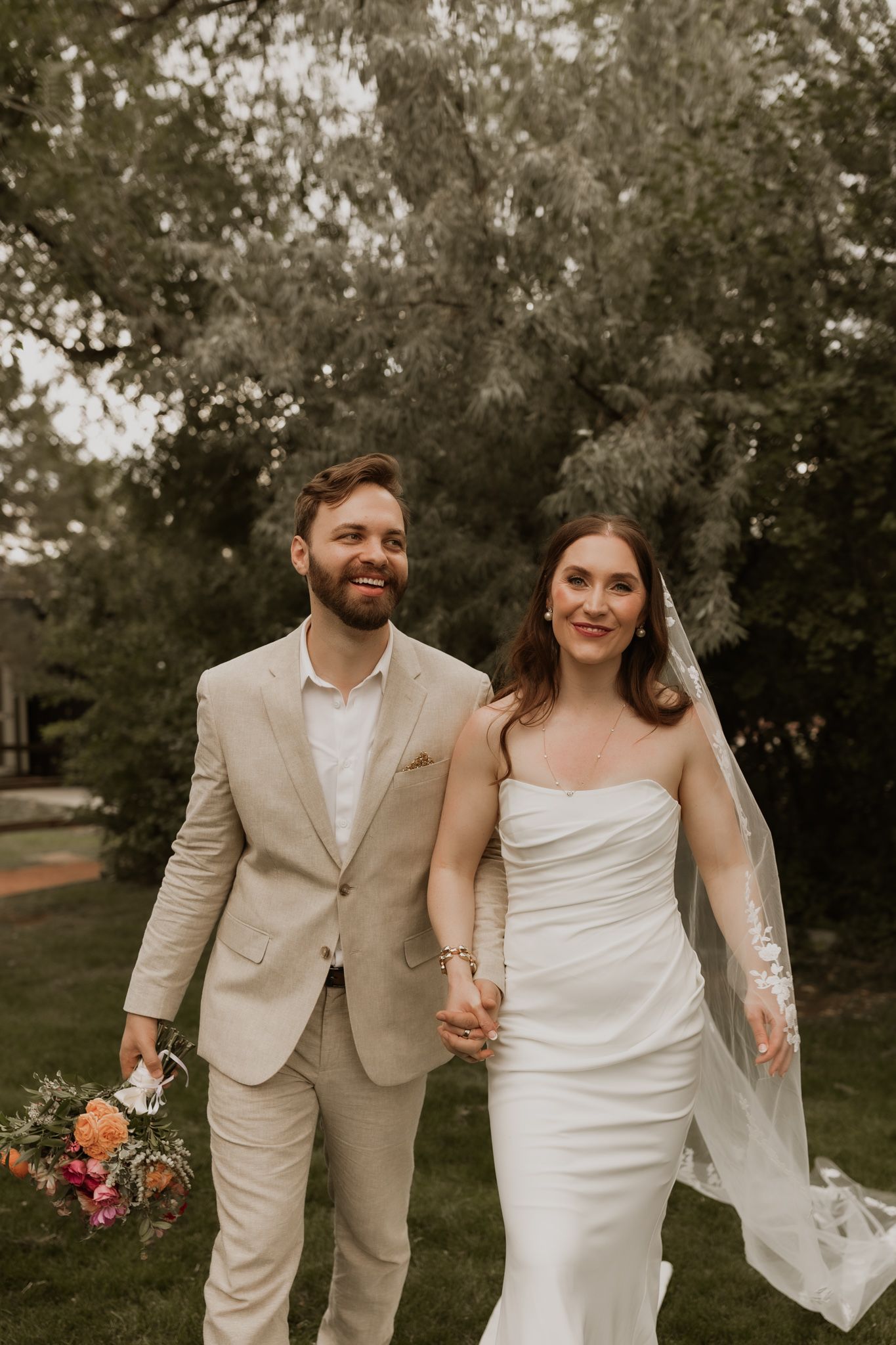 Joyful wedding couple walking together outdoors in coordinated beige and white attire.